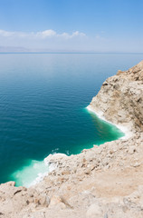 Overlooking the Dead Sea with its turquoise and aquamarine water and rugged, salt crusted bank. The horizon is hazy with blue sky and clouds above. 
