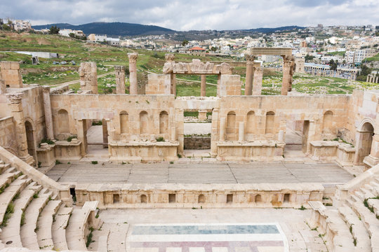Roman Theater at Jerash, Jordan with view of Jerash city behind the stage in the background and cloudy sky above. The floor of the theater has a mosaic floor.