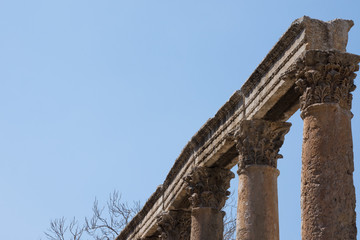 Row of stone columns topped with ornately carved capitals and stone lintel or beams. Ancient ruins are located in Amman Jordan and have blue, cloudless sky in the background. Photographed at an angle.