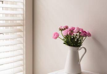 Pink ranunculus in white jug on table next to window with blinds