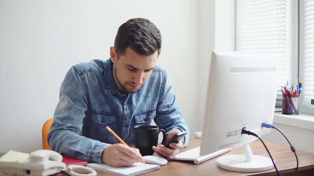 Young businessman sitting by the computer in stylish modern office and taking notes using his pencil and phone. Computer, phone and cup on the table. Shot in 4k