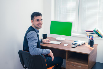 Happy office worker smiling and looking at the camera. He sits at computer with Chromakey. Office concept with working environment. Copyspace on screen