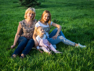 Happy family of Grandmother, Mother and daughter smiling at the park having picnic. They are sitting and resting on green lawn with grass