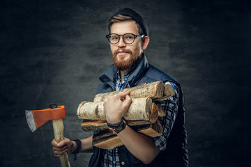 Bearded male in eyeglasses dressed in fleece shirt holds firewoods and axe.