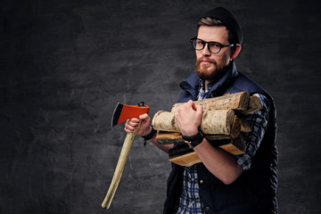 Bearded male in eyeglasses dressed in fleece shirt holds firewoods and axe.