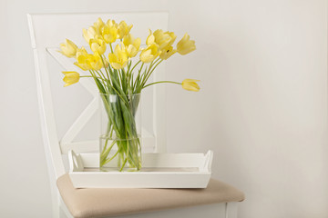 Yellow tulips in vase on white tray on the chair.