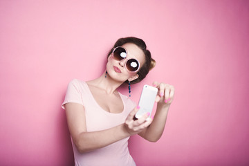 close-up portrait of young beautiful slim sexy young blonde woman in a retro pin-up style on pink background in studio wearing sunglasses smiling and posing selfi doing on your phone