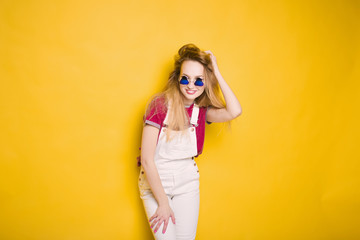 close-up portrait of a beautiful young hipster lifestyle slim blond girl in fashionable sunglasses on a yellow background in the studio in a white suit and shirt smiling and posing