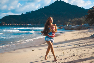 Fototapeta premium close-up portrait of a beautiful young brunette girl with long hair on a background of blue sea with waves and sky with clouds on a sunny day, lifestyle, posing and smiling, wind