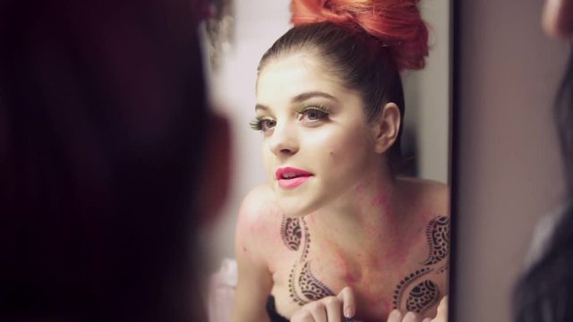 Beautiful young model with false eyelashes looking in the mirror at herself while preparing to the fashion show. Makeup artist standing behind. Slowmotion shot