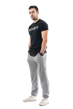 Profile View Of Serious Muscular Bouncer Or Bodyguard Looking At Camera Skeptically. Full Body Length Portrait Isolated On White Studio Background. 