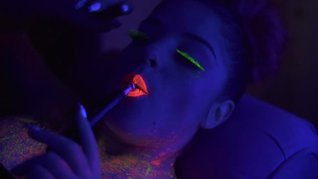 Portrait of a young beautiful woman with fluorescent makeup inhaling hookah under UV black light. Sexy girl smoking shisha lying on sofa. Slowmotion shot