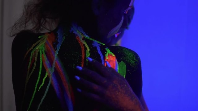 Closeup view of two beautiful girls with fluorescent makeup and body art under UV black light petting each other and hugging. Slowmotion shot