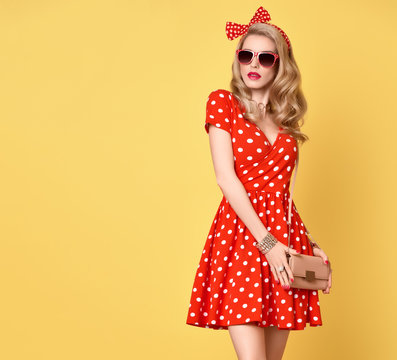 Fashion PinUp Model Girl in Red Polka Dots Summer Dress. Stylish Curly hairstyle, Trendy Clutch, fashion Headband, Sunglasses. Beauty Blond Woman in fashion pose. Glamour Playful Sexy Lady on Yellow