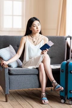 asian woman traveler sitting on sofa with suitcase and passport, packing luggage
