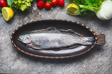 Raw fresh whole trout in baking dish with vegetables cooking  ingredients on gray concrete background , top view.  Healthy Food concept