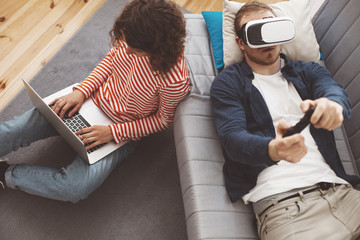 Young couple using electronic devices. Man playing breathtaking computer game with VR headset. Woman typing on laptop. Modern family life