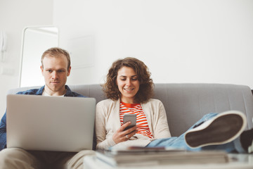 Young family spending time with electronic devices. Man using laptop and smiling woman typing message with mobile phone. Sitting together on the sofa