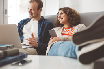 Fototapeta premium Pretty family spending time with electronic devices. Man using laptop and smiling woman typing message with mobile phone. Sitting together on the sofa