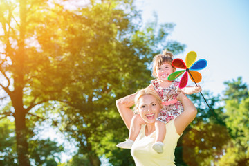 Smiling mother holding her little daughter on the shoulders, little girl has a pinwheel toy.Lens flare, copy space