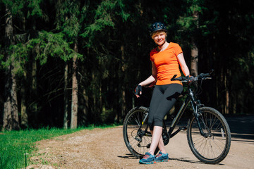 Young pretty woman in helmet and orange shirt with bicycle resting on the forest road after ride. Healthy lifestyle concept. Sports Girl with bike.