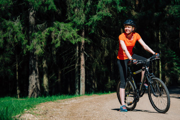 Obraz premium Young pretty woman in helmet and orange shirt sitting on bicycle on the forest road. Healthy lifestyle concept. Sports Girl with bike.