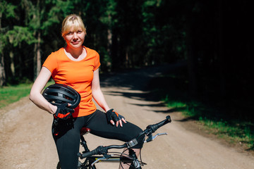 Fototapeta na wymiar Young pretty woman in orange shirt and with helmet in hand sitting on bicycle on the forest road. Healthy lifestyle concept. Sports Girl with bike.