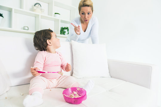 Mother scolding her little baby girl while she eating a cake on  sofa and pieces of cake falling down all around.Copy space, shallow doff
