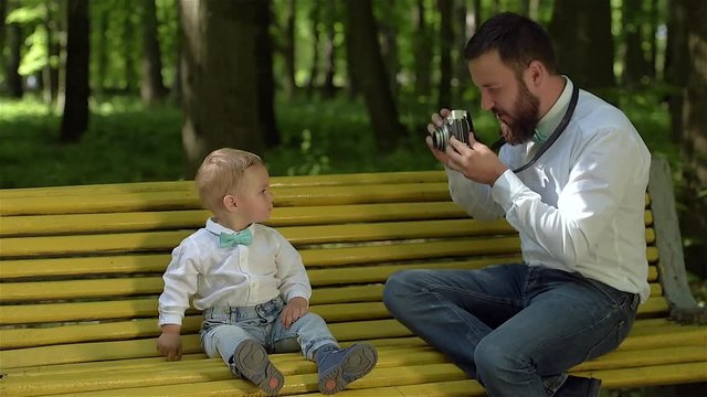 A young father with a beard sits on a yellow bench in the park and takes pictures of his young son. Father's Day.
