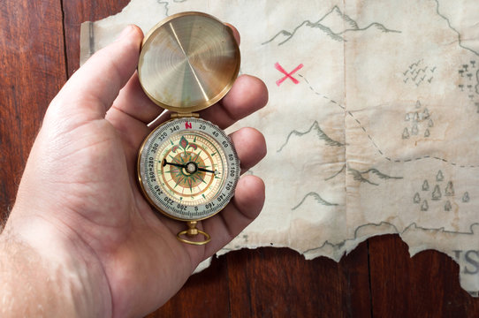 Man holds compass above fake pirate map of abstract island with red cross of place where is treasure