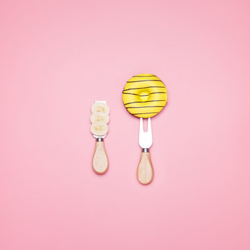 Top view of a yellow donut on a fork and a sliced banana on a pastel pink background. Minimal design.