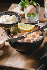 Black bowl of salmon pate with red caviar served with butter, sliced bread, capers, vintage knife, verrines and herbs on wooden serving board, textile linen over brown texture background. Close up