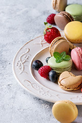 Variety of colorful french sweet dessert macaron macaroons with different fillings served on white vintage plate with spring flowers and berries over gray texture background. Close up with space