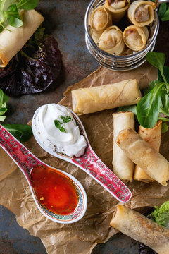 Fried spring rolls with red and white sauces in china spoons, served on crumpled paper and in fry basket with green salad and wooden chopsticks over old metal texture background. Top view. Asian food