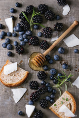 Berries blackberry and blueberry, honey on dipper, rosemary, sliced goat cheese with bread served over gray metal texture background. Summer sandwich. Top view