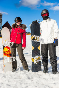 Stylish couple in ski suits, helmets and ski goggles standing with snowboards in a ski resor