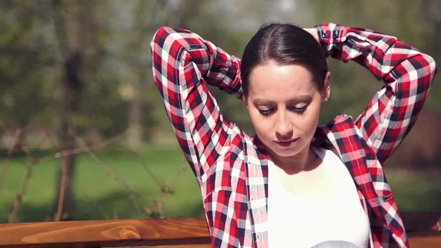 Beautiful young girl combing her long hair by hands. Young femail sitting on a bench in the park and relaxing in the sun. HD 1080p.