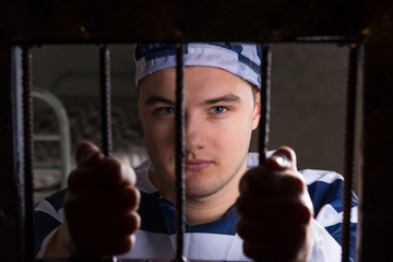 View through iron door with prison bars on young male prisoner holding bars in a jail cell