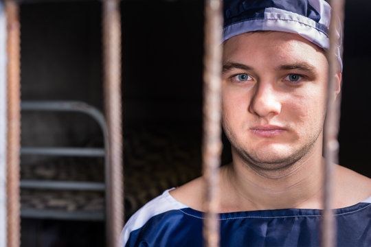 Portrait of young male prisoner standing behind a prison grid in a jail cell