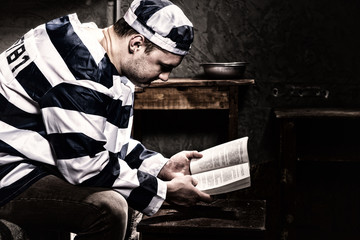 Male prisoner wearing prison uniform reading a book or a bible while sitting on a bed in a prison...