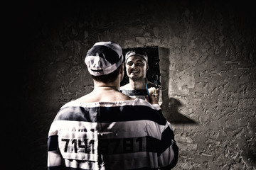 Prisoner standing with toothbrush and looking at his reflection in the mirror in a prison cell