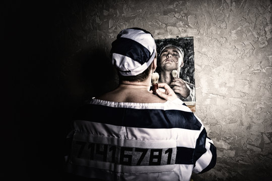 Male prisoner standing with shave brush and looking at his reflection in the mirror in a prison cell