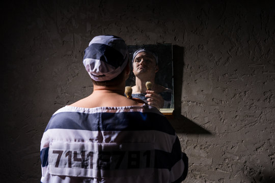 Prisoner standing with shave brush and looking at his reflection in the mirror in a prison cell