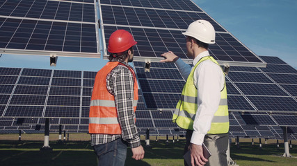 Technicians in long sleeve shirts, reflective vests and hard hats discussing something about solar panel power arrays outside