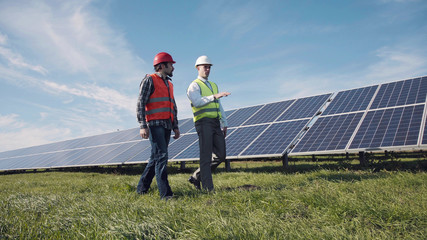 Two male electrician workers in reflective vests and hard hats walking in between long rows of photovoltaic solar panels and talking about installation of new solar panels.