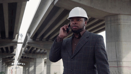 Smiling african man wearing white protective helmet talking on mobile phone while standing under the overpass construction and then looking at camera