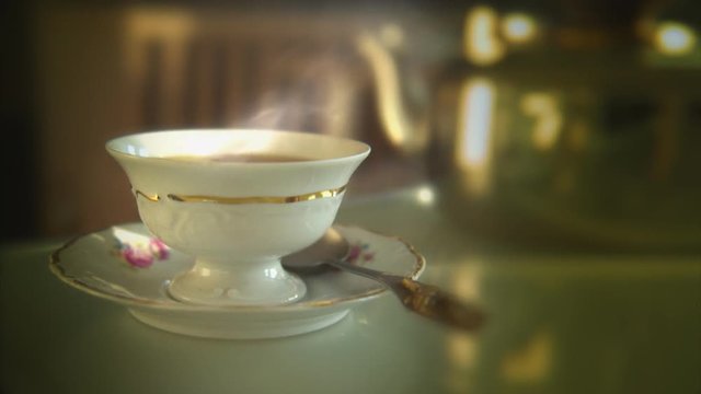 Vintage porcelain cup full of hot tea on a glass tabletop. Defocused metal pot in the background.
