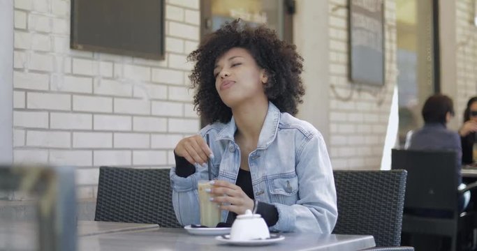 Charming young black woman with short curly hair wearing denim jacket and holding straw in glass while looking away.