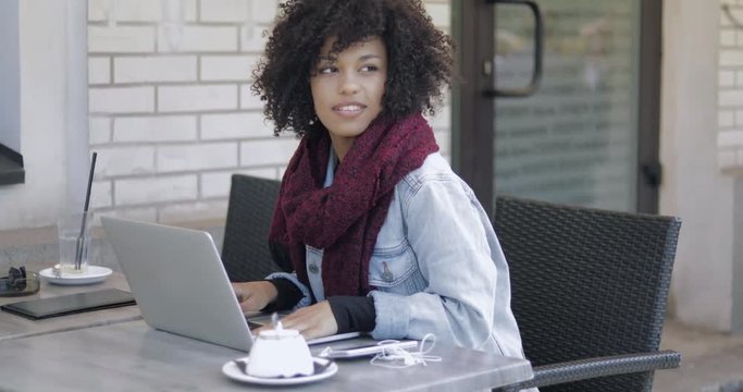 Beautifulblack woman with short curly hair sitting at tablein outside cafe with laptop and typing.
