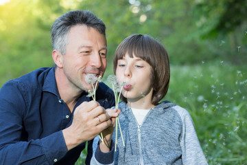Man with his son blowing dandelions over blurred green grass, summer nature outdoor